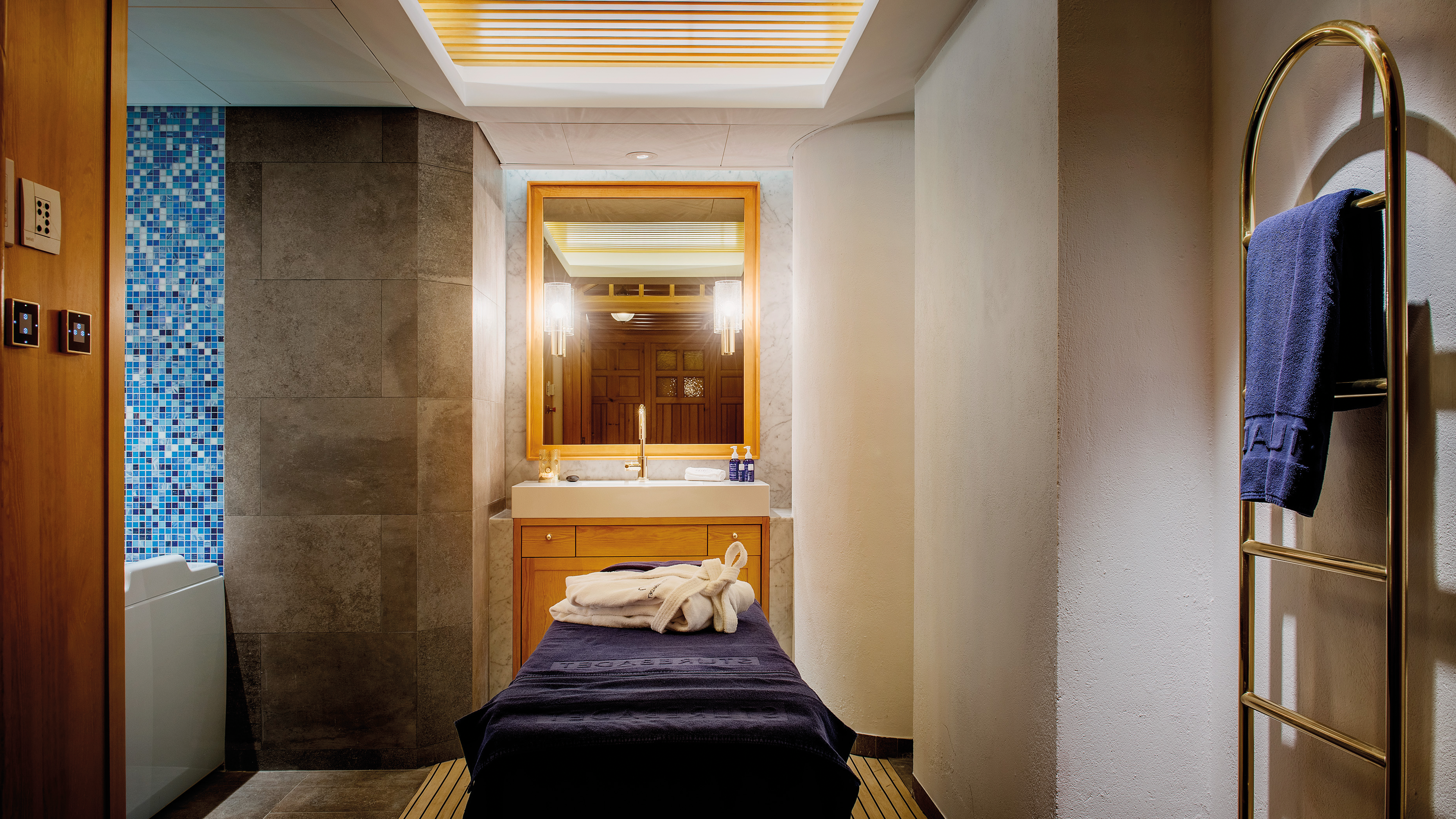 Massagerum på Sturebadet med en mässings handdukstork, spegel lampetter på vardera sida och en massagesäng med blå handduk i mitten. 
