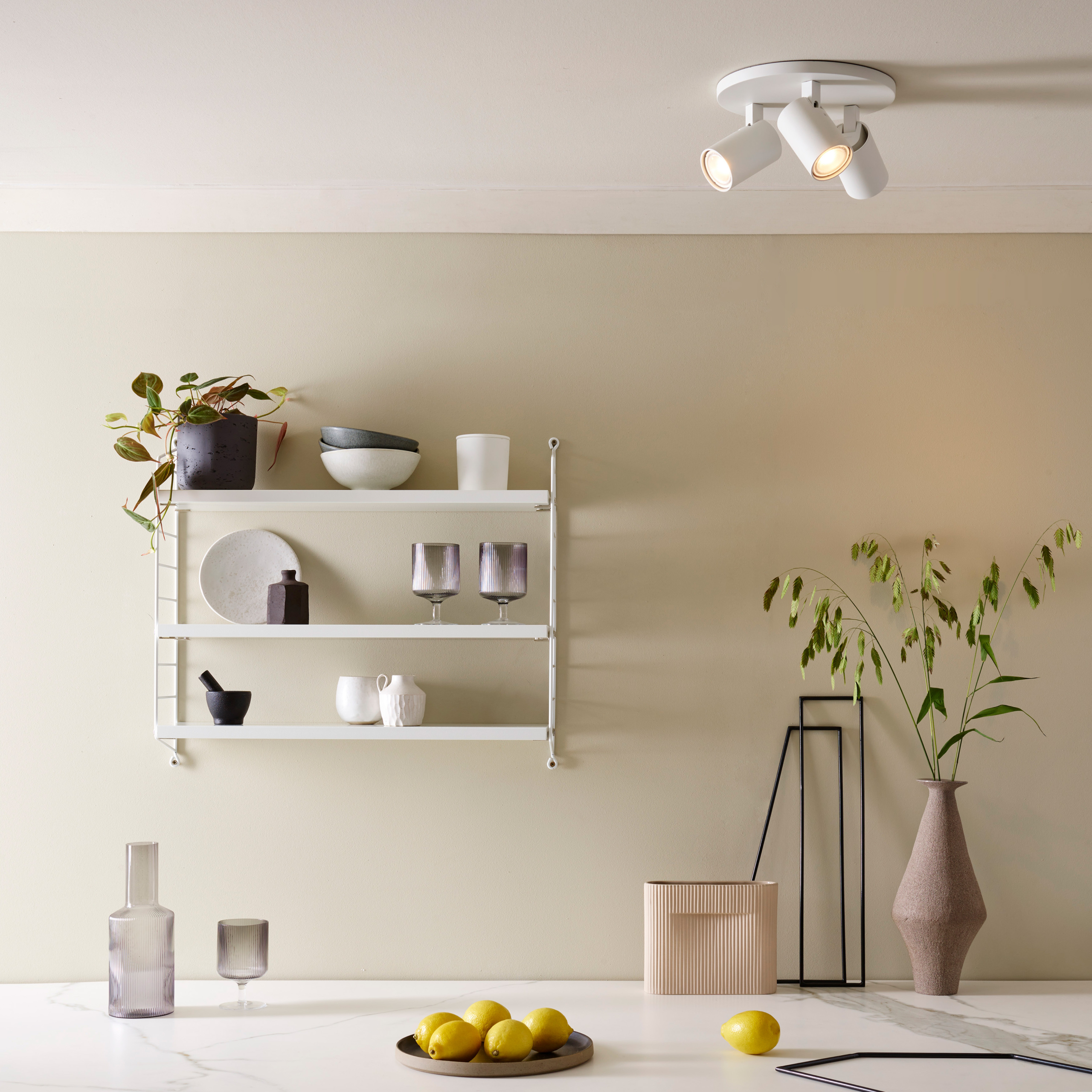 Ascoli Triple Round taklampa med 3 spotlights på en rund bas. under lampan på väggen finns en hylla med glas och ett bord med en skål med citroner.