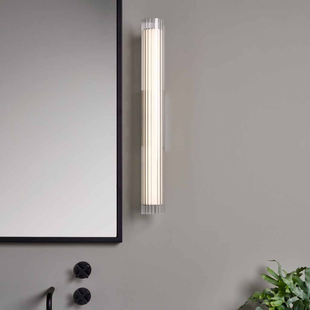 io 665 vägglampa i glas i en cyliderform bredvid en spegel.
