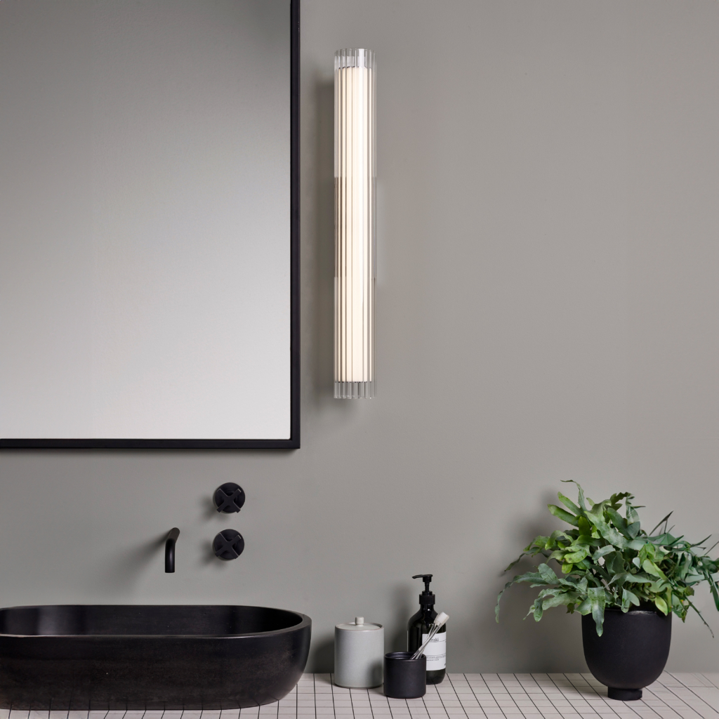 io 665 vägglampa i glas i en cyliderform bredvid en spegel.