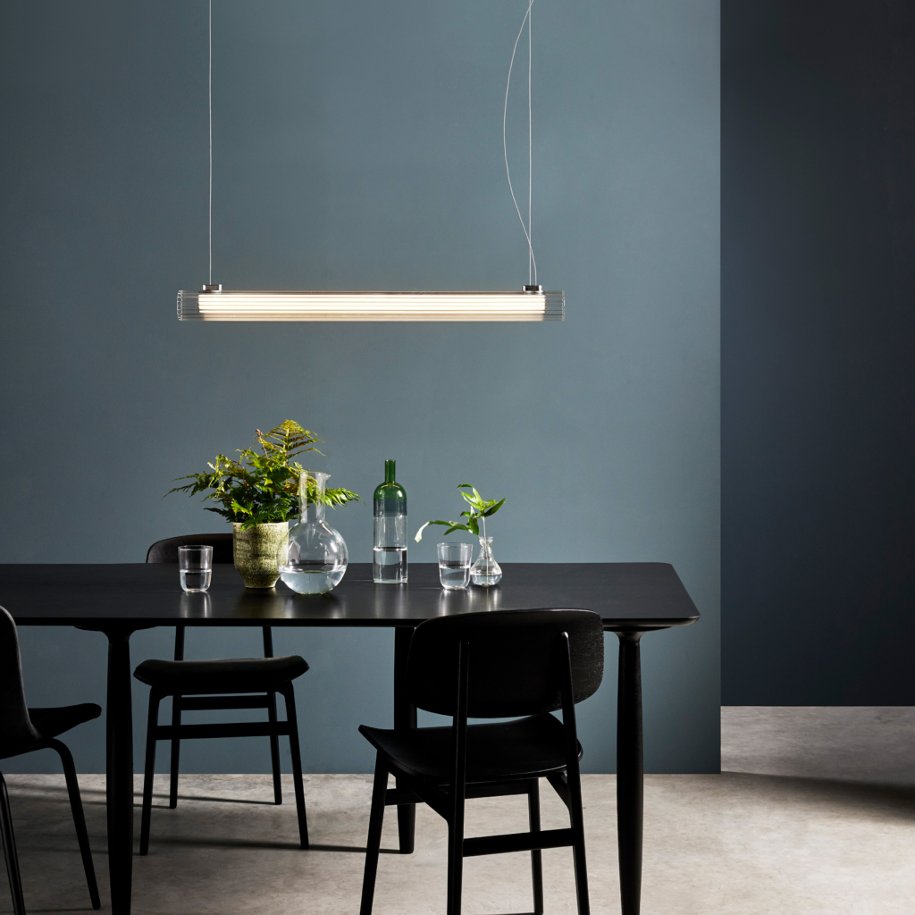 io Pendant 1000 taklampa, är en cylinderformad lampa som hänger från taket i två stållinor. Under lampan står et bord med fyra stolar. på bordet fins karaff och en vas med blommor.