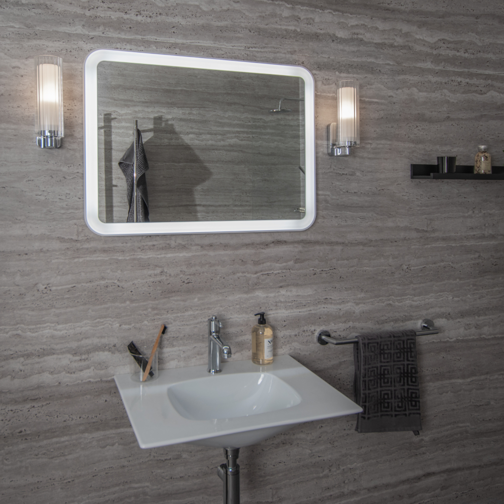 Elegant rektangulär väggspegel sittandes på en trävägg med ett vitt tvättställ under och e lampa på vardera sida om spegeln.