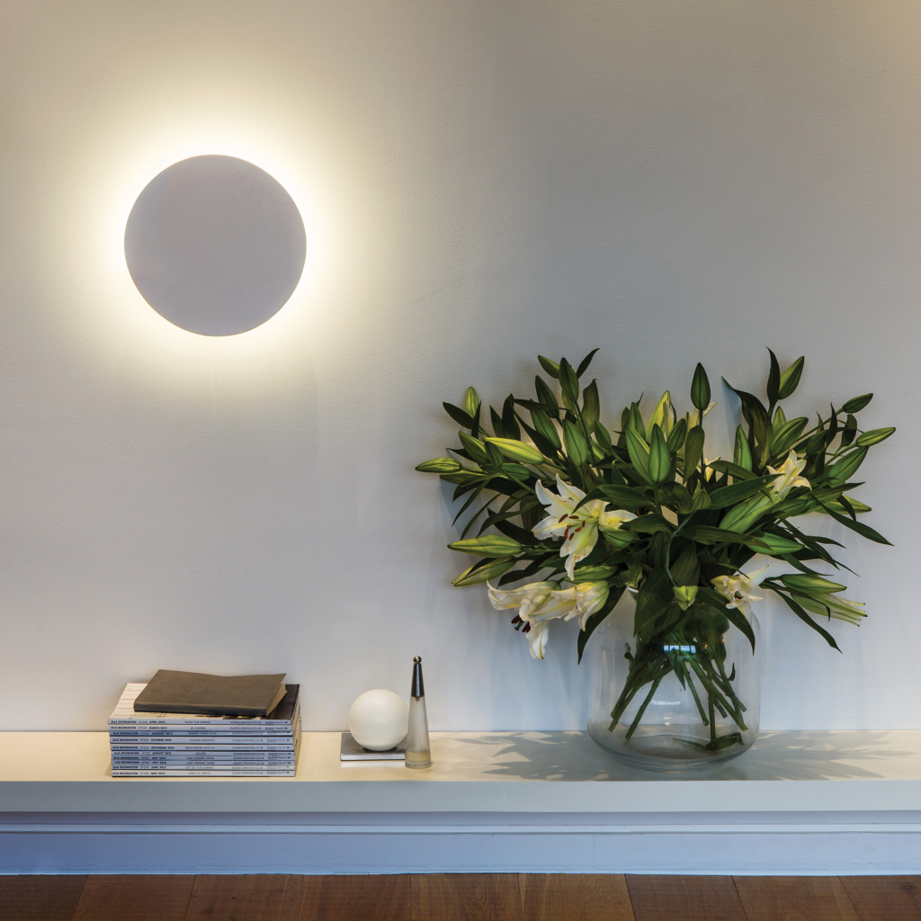 Eclipse Round 250 LED vägglampa sitter på en vägg vid ett bord med en bukett blommor och böcker.